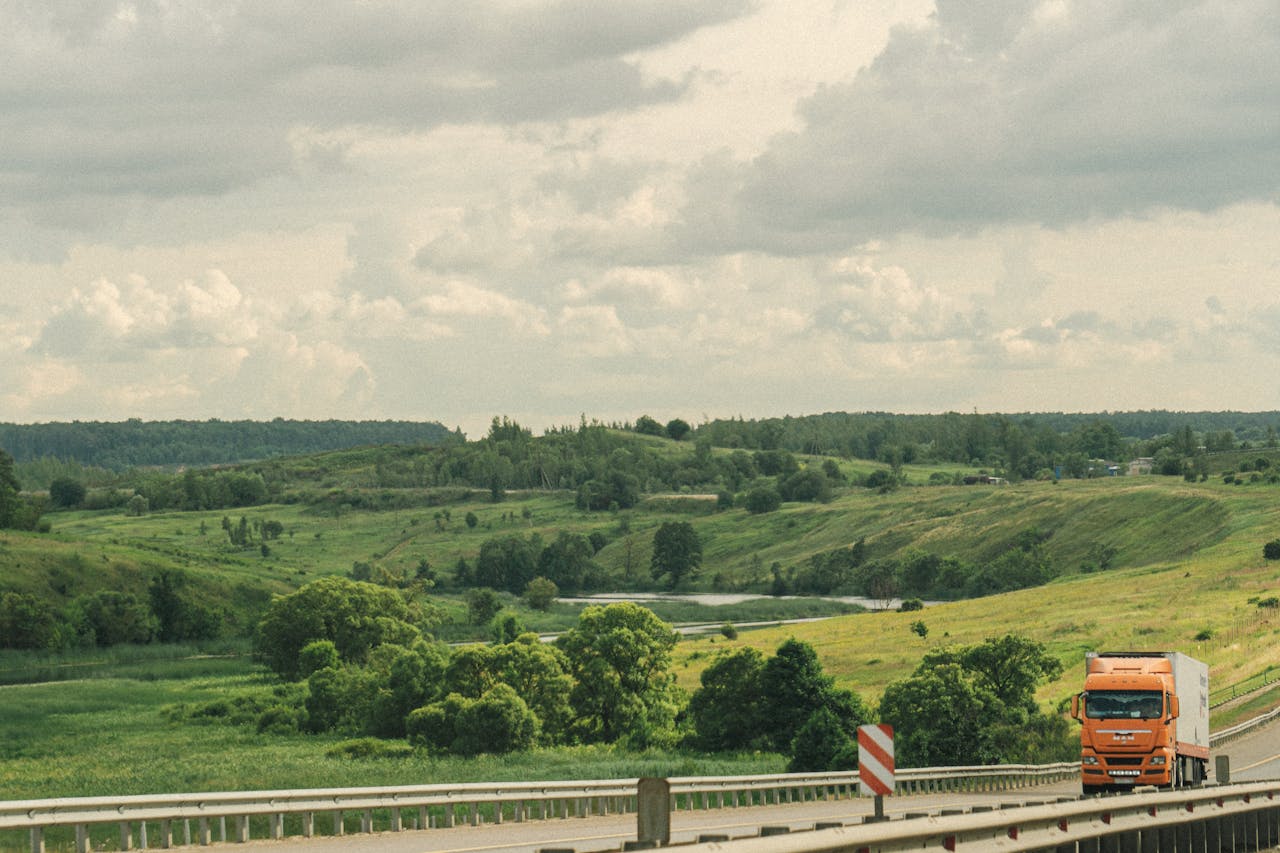 An orange truck drives along a highway through a lush, green, hilly countryside under a cloudy sky.